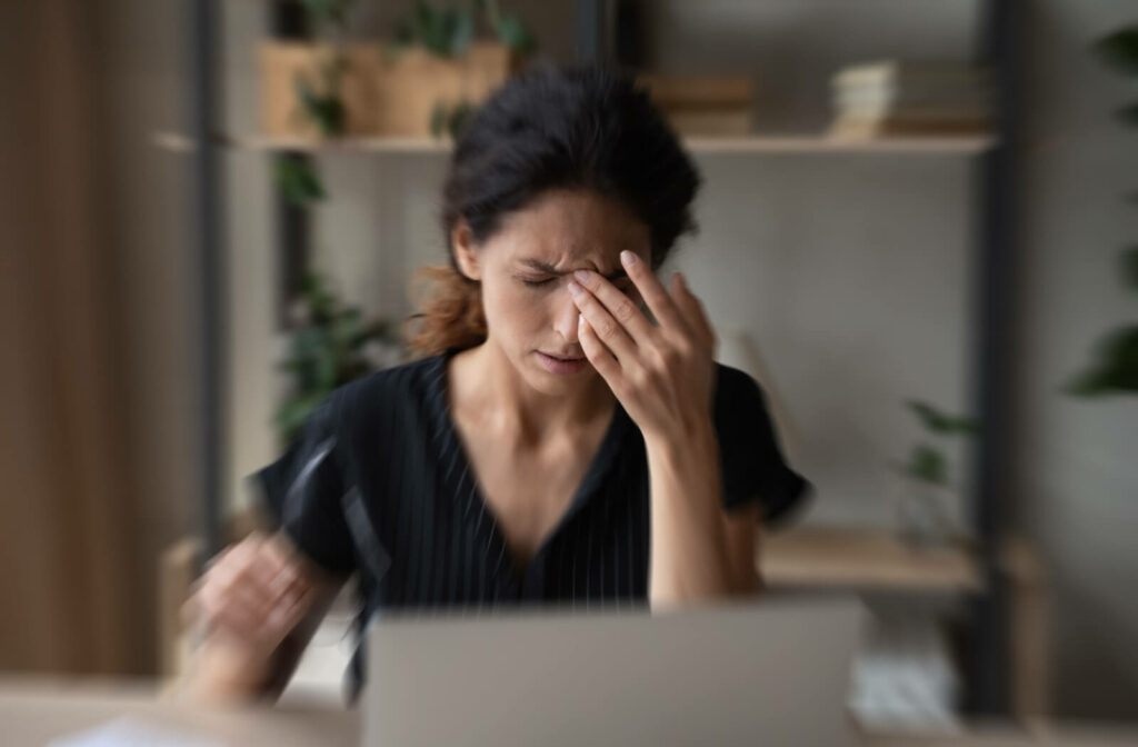 A woman sitting in front of her computer took off her glasses and touching her forehead, experiencing dizziness with her new eyeglasses.