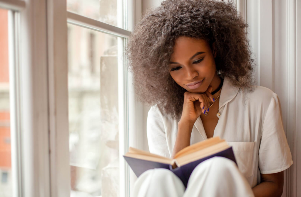 Young woman with curly hair enjoying a book while sitting on a window sill.