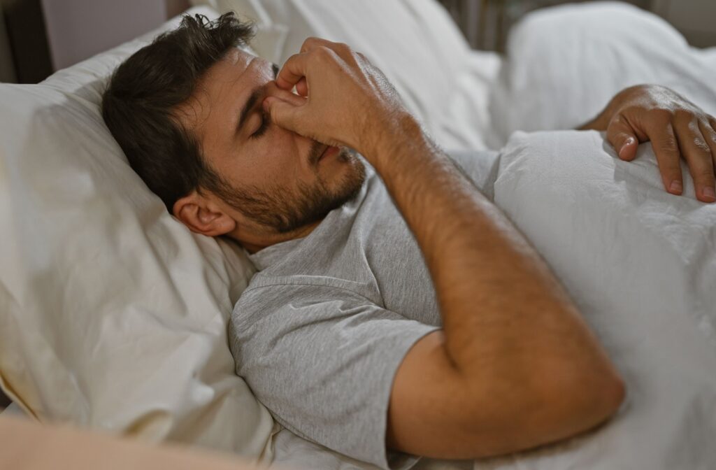 A young adult man lying in bed indoors, covering his eyes with his hand, showing a sense of discomfort