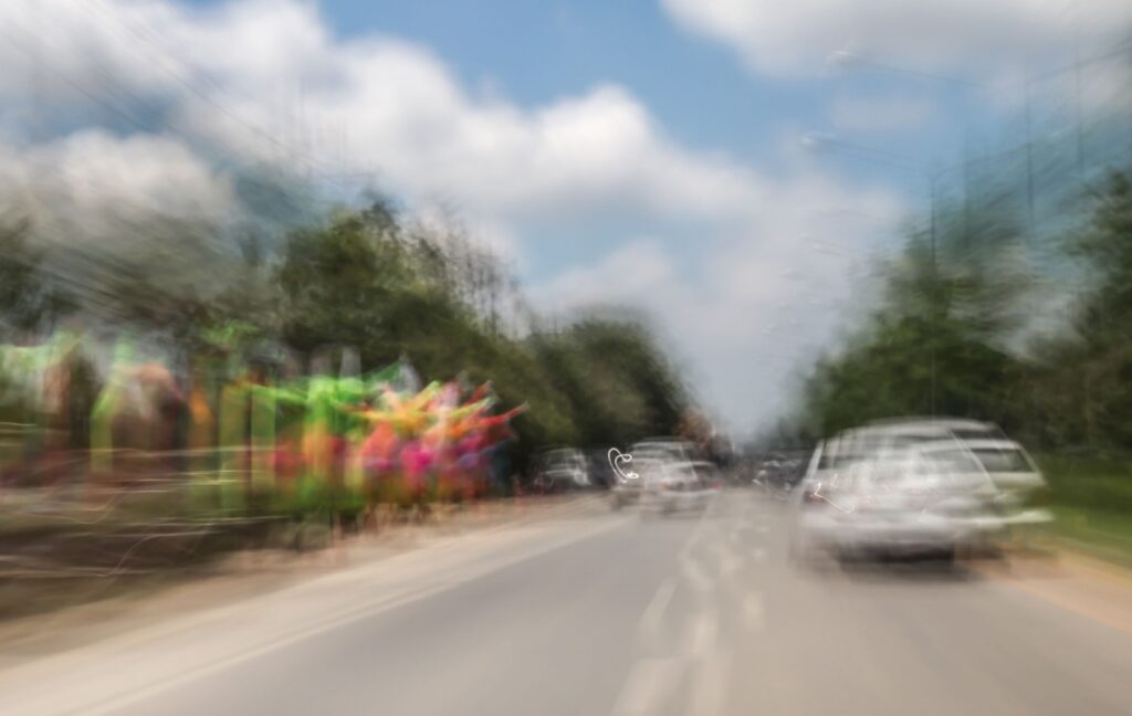 The image shows a scene of a street with cars and lush green trees, blurred by double vision.