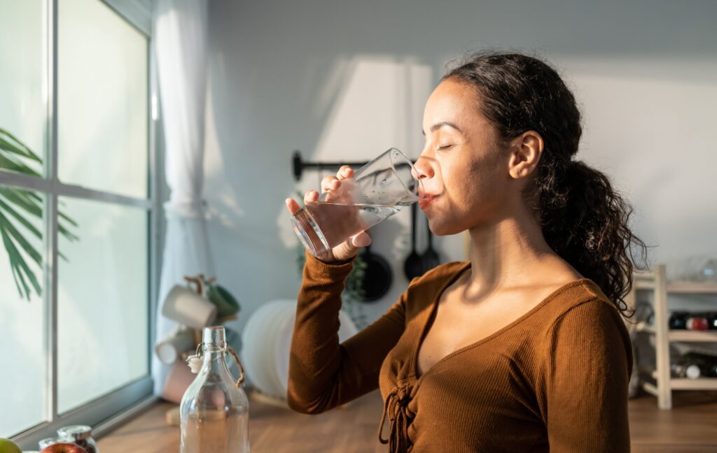 A person stands in front of a bright window drinking a large glass of water to stay hydrated.