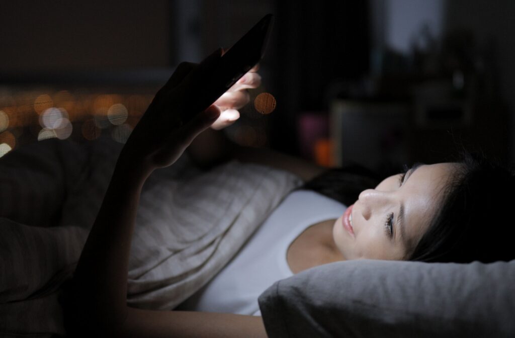 A person lies in bed at night, holding and looking at a smartphone in a dark room.