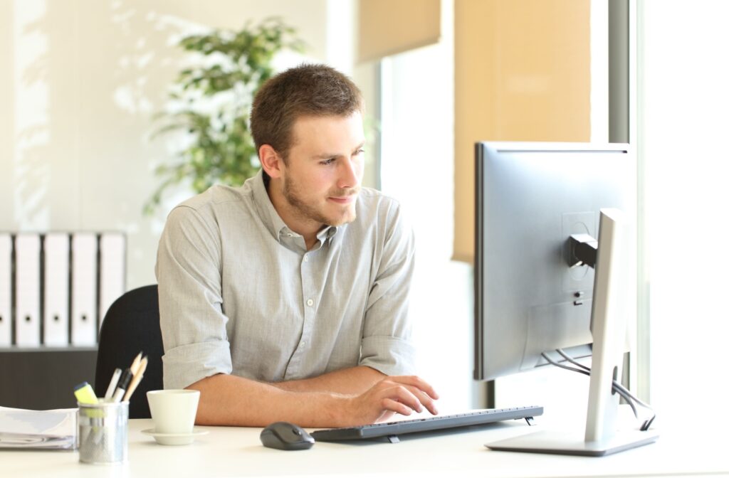 A person sits at a desk, focused on a computer screen in a bright office setting.