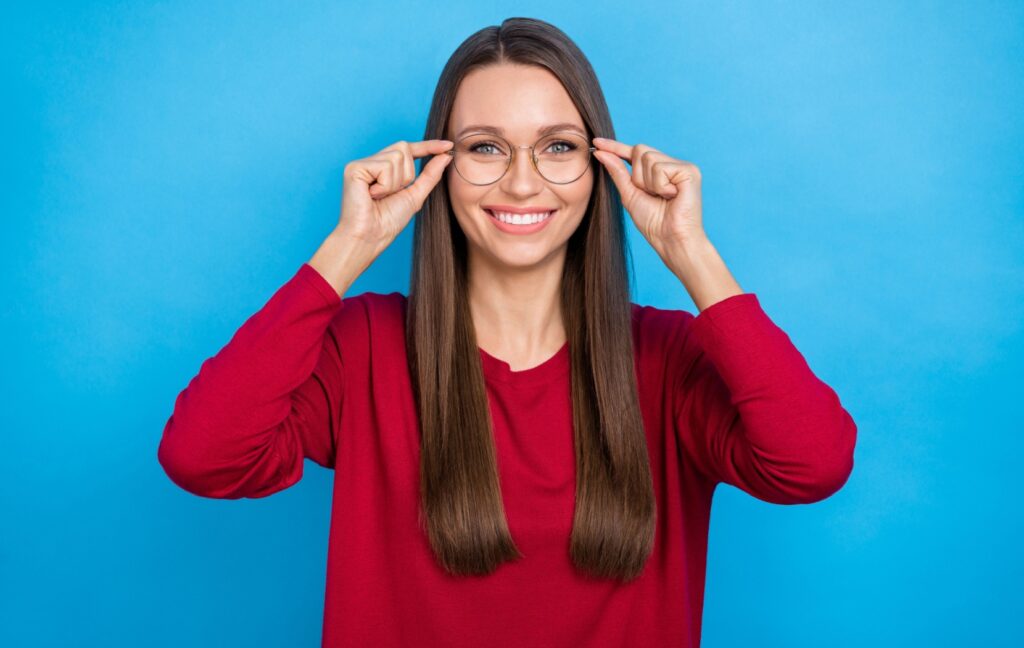 A young adult slides a pair of brand new glasses onto their face while smiling in front of a blue background.