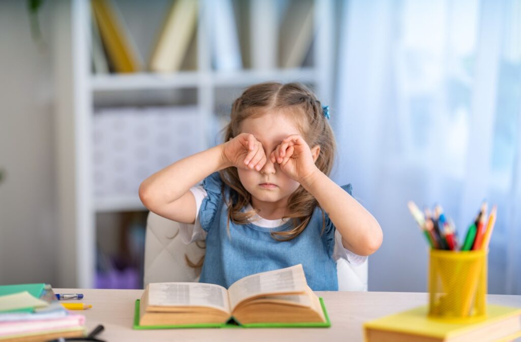 A young child sitting at a desk with a book open in front of them, rubbing their eyes due to vision problems.